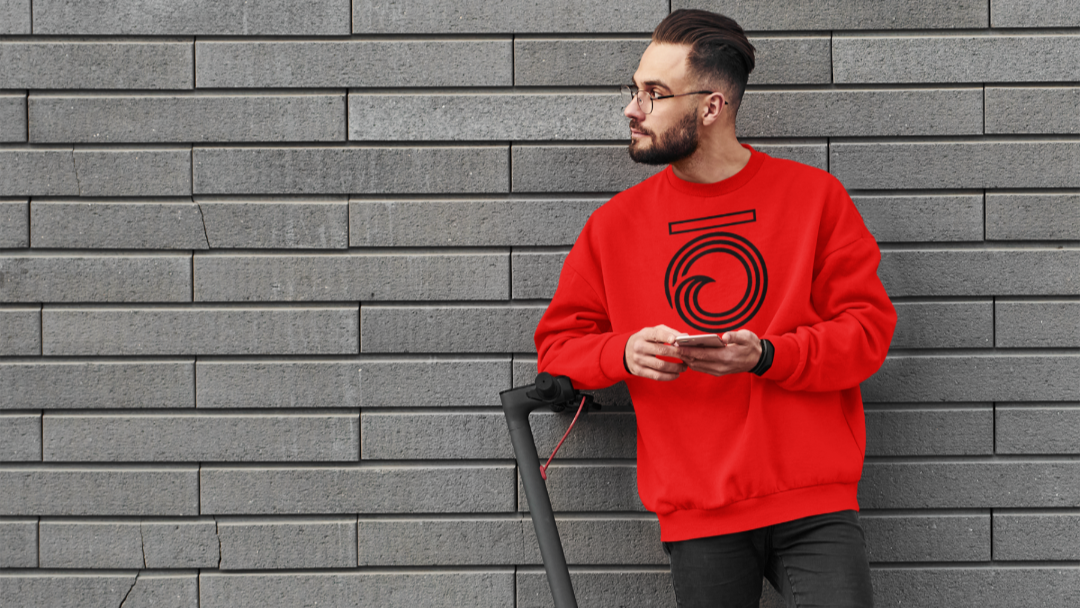 Man in a red sweatshirt with a logo standing next to a scooter against a gray brick wall.